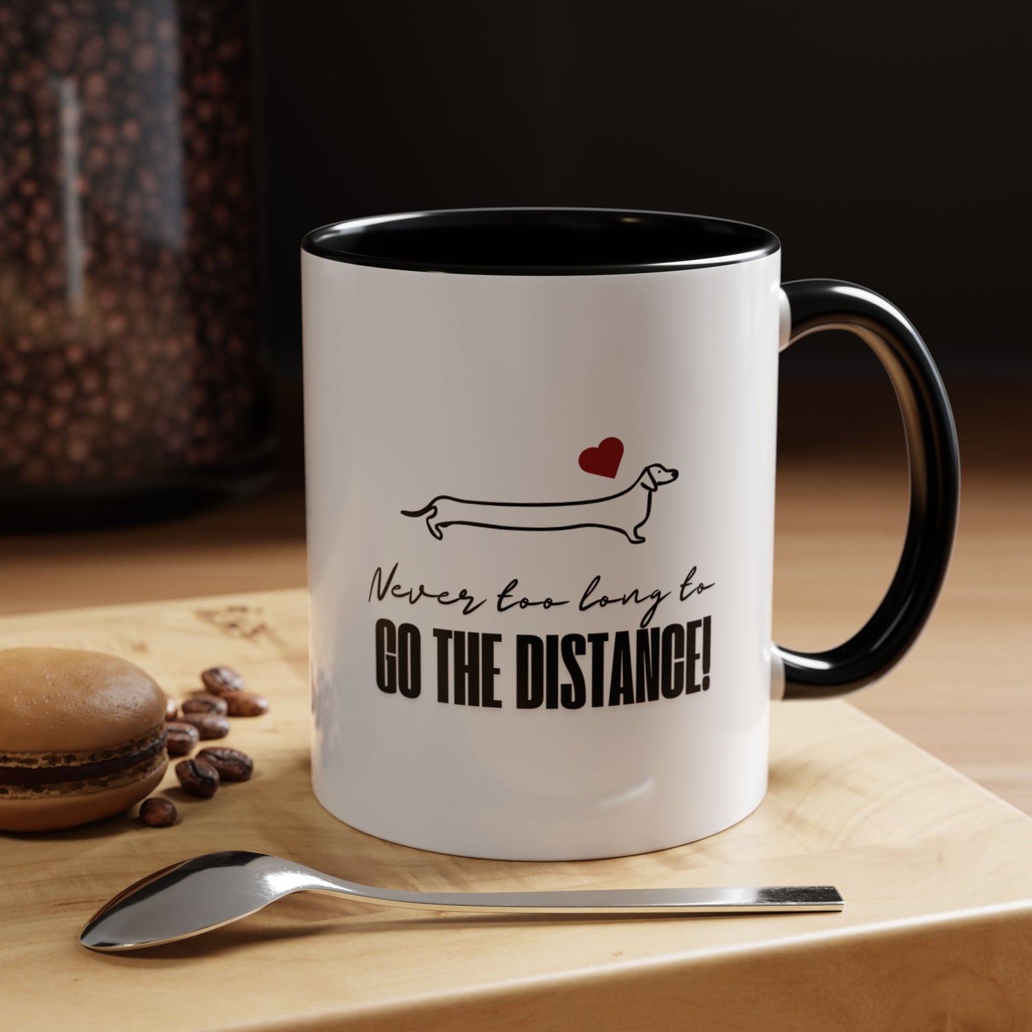 Dachshund coffee mug on a wooden table with spoon, coffee beans, and a macaron, featuring a minimalist Dachshund design, red heart, and the phrase “Never too long to GO THE DISTANCE!”