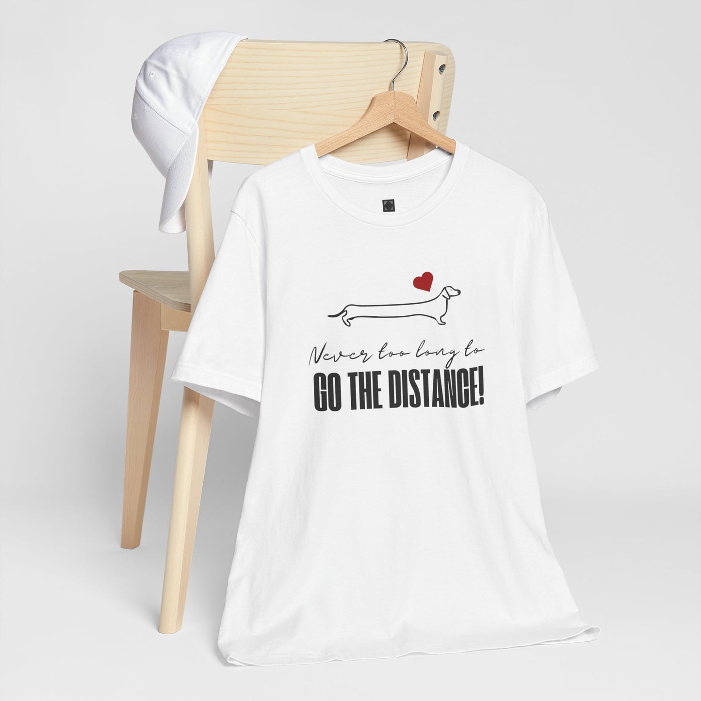 White Dachshund t-shirt hanging over a chair, featuring a minimalist Dachshund graphic, red heart, and the phrase “Never too long to GO THE DISTANCE!