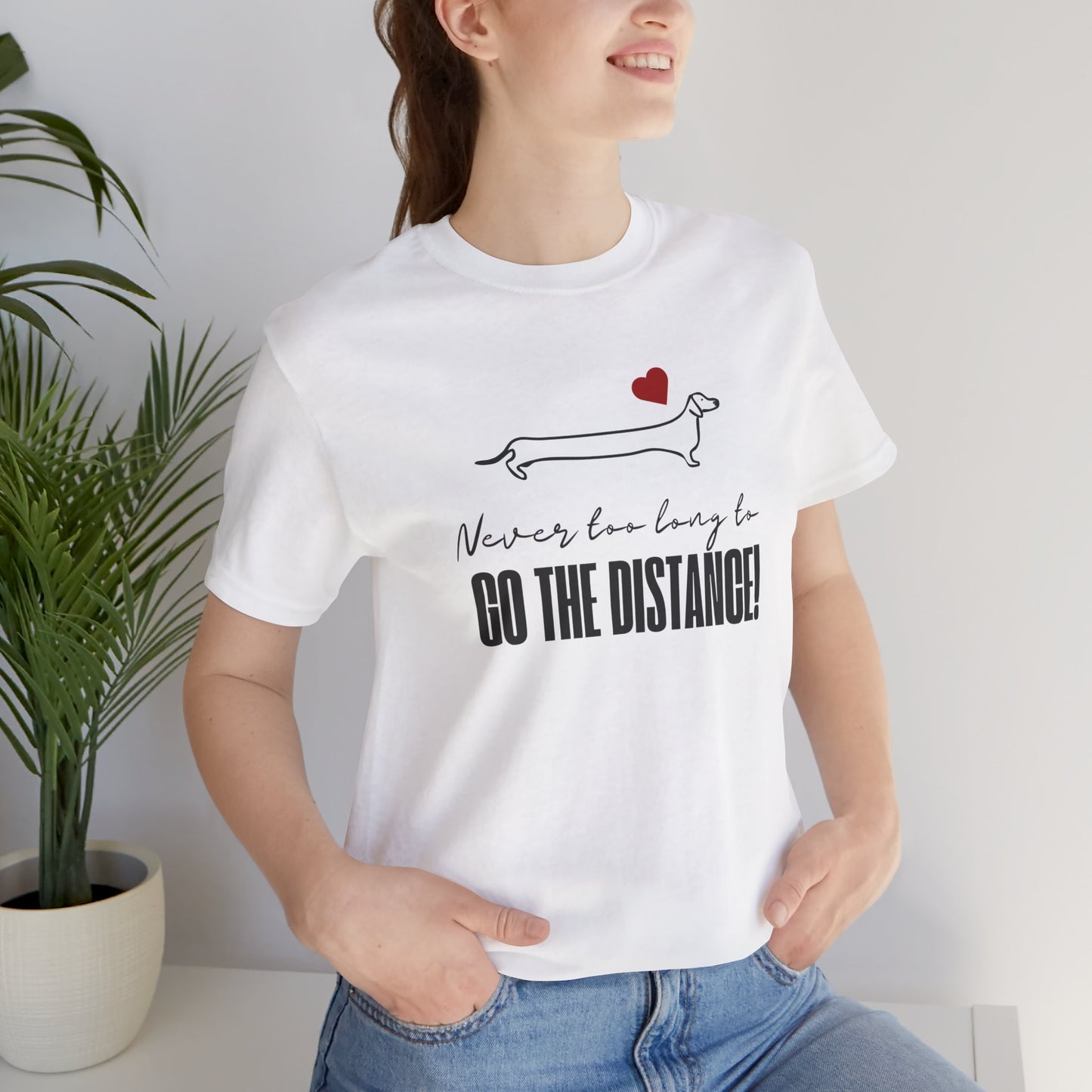Woman wearing a white motivational dachshund t-shirt that says "Never too long to GO THE DISTANCE!" with a minimalist dachshund graphic and red heart above it.