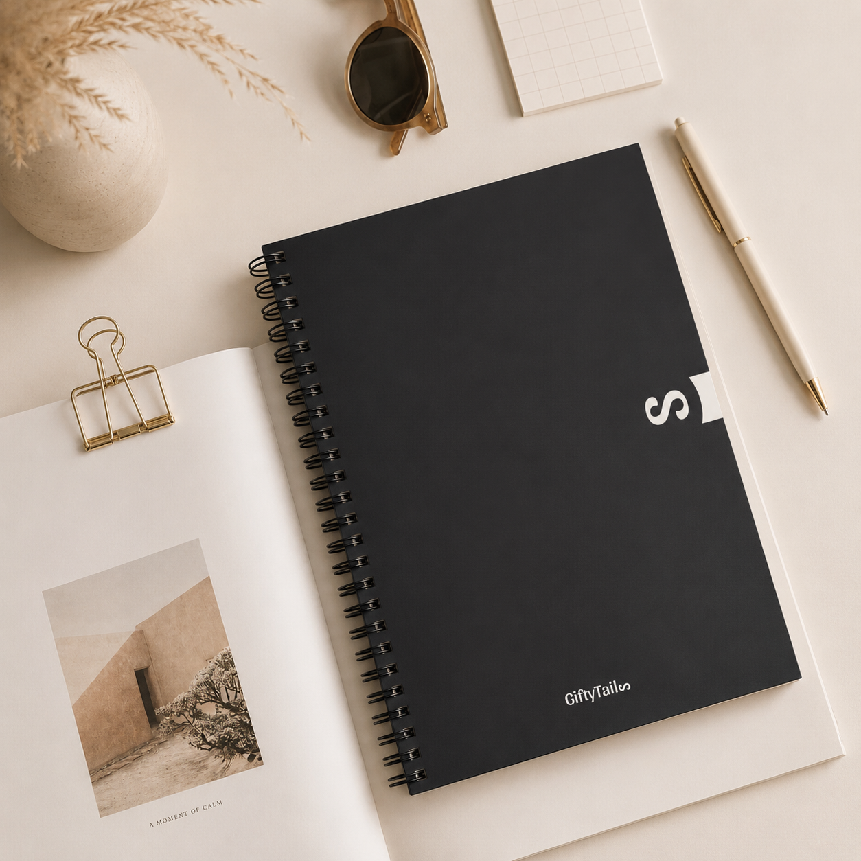 Minimal black spiral notebook flat lay styled on a soft neutral desk with sunglasses, beige pen, dried pampas grass, and clean aesthetic stationery accessories.
