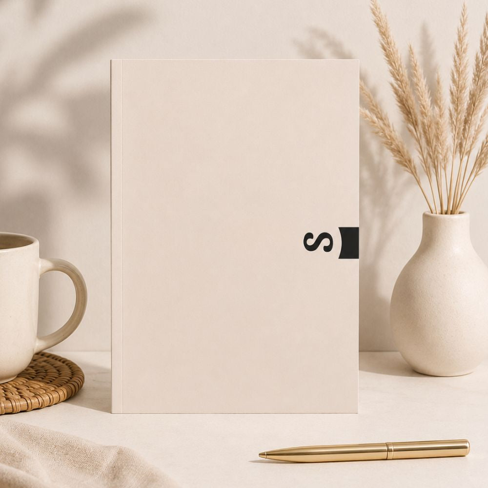 Minimal beige A5 softcover planner standing on a neutral desk with a ceramic mug, dried pampas grass vase, and gold pen in a calm aesthetic workspace setting.