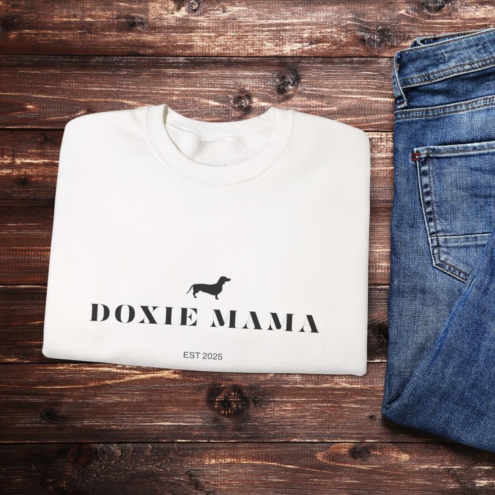 Folded white Doxie Mama crewneck sweatshirt with dachshund graphic, laid next to jeans on wood background